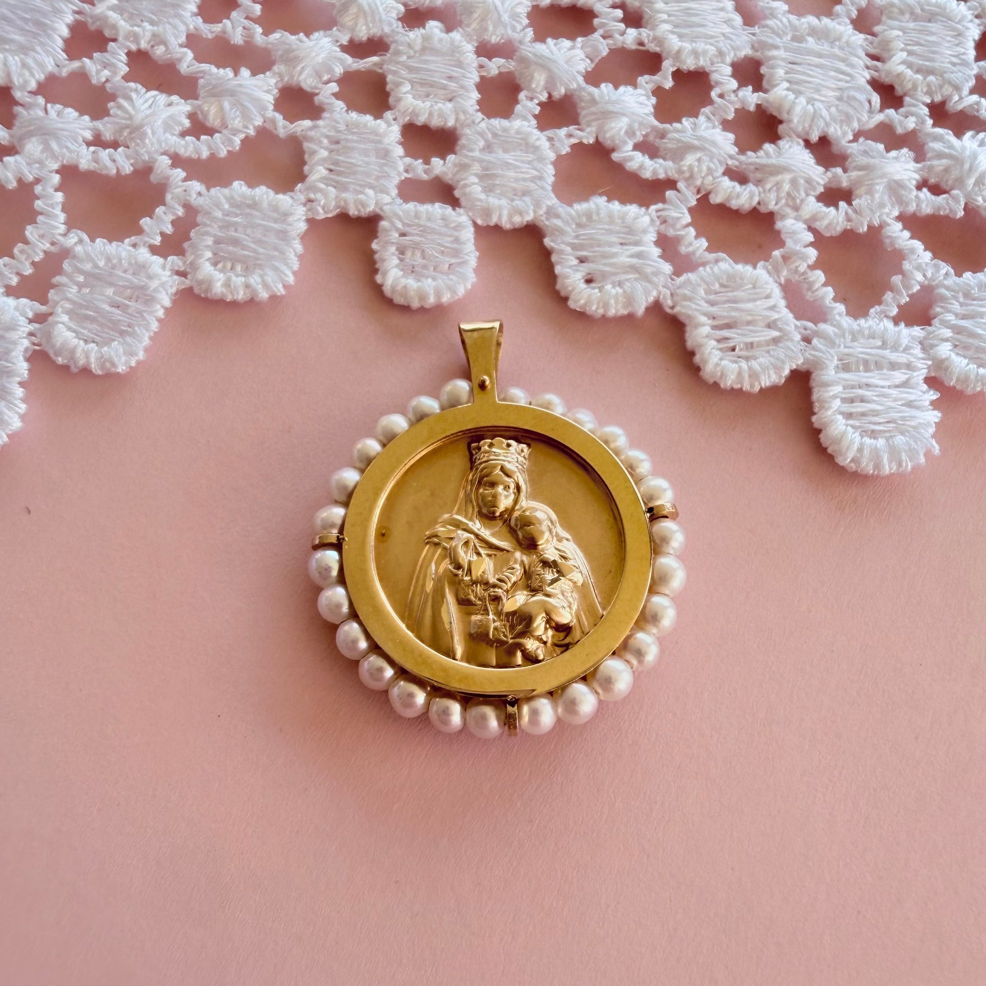 A 14k gold  religious pendant featuring a carved  Our Lady of Mt. Carmel and the Sacred Heart, surrounded by a border of small white pearls, displayed on a soft pink surface with a white lace cloth in the background.
