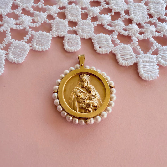 A 14k gold  religious pendant featuring a carved  Our Lady of Mt. Carmel and the Sacred Heart, surrounded by a border of small white pearls, displayed on a soft pink surface with a white lace cloth in the background.